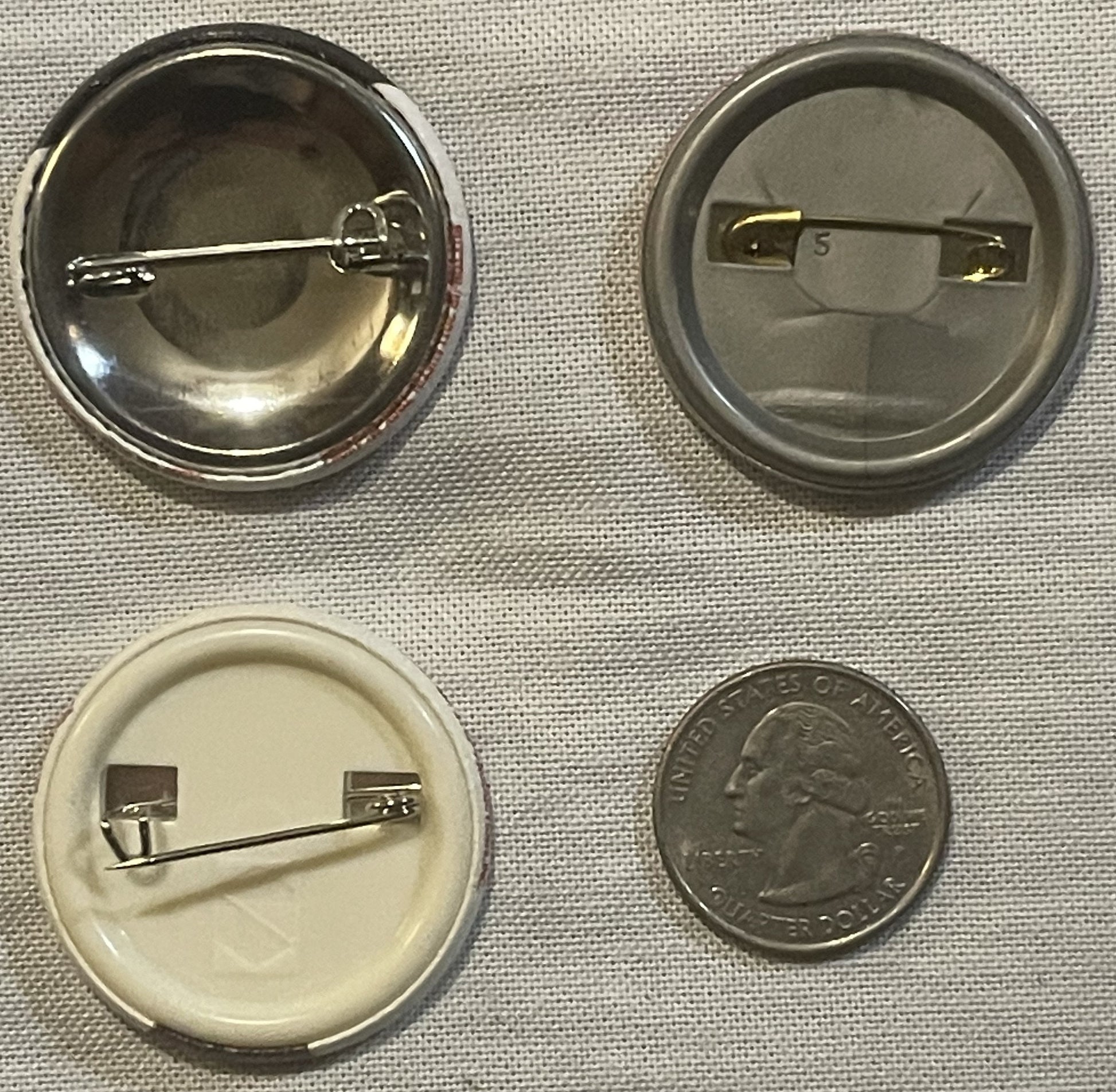 Four button pin backs shown front and back with a quarter for scale, including two metal pin mechanisms and two plastic backi