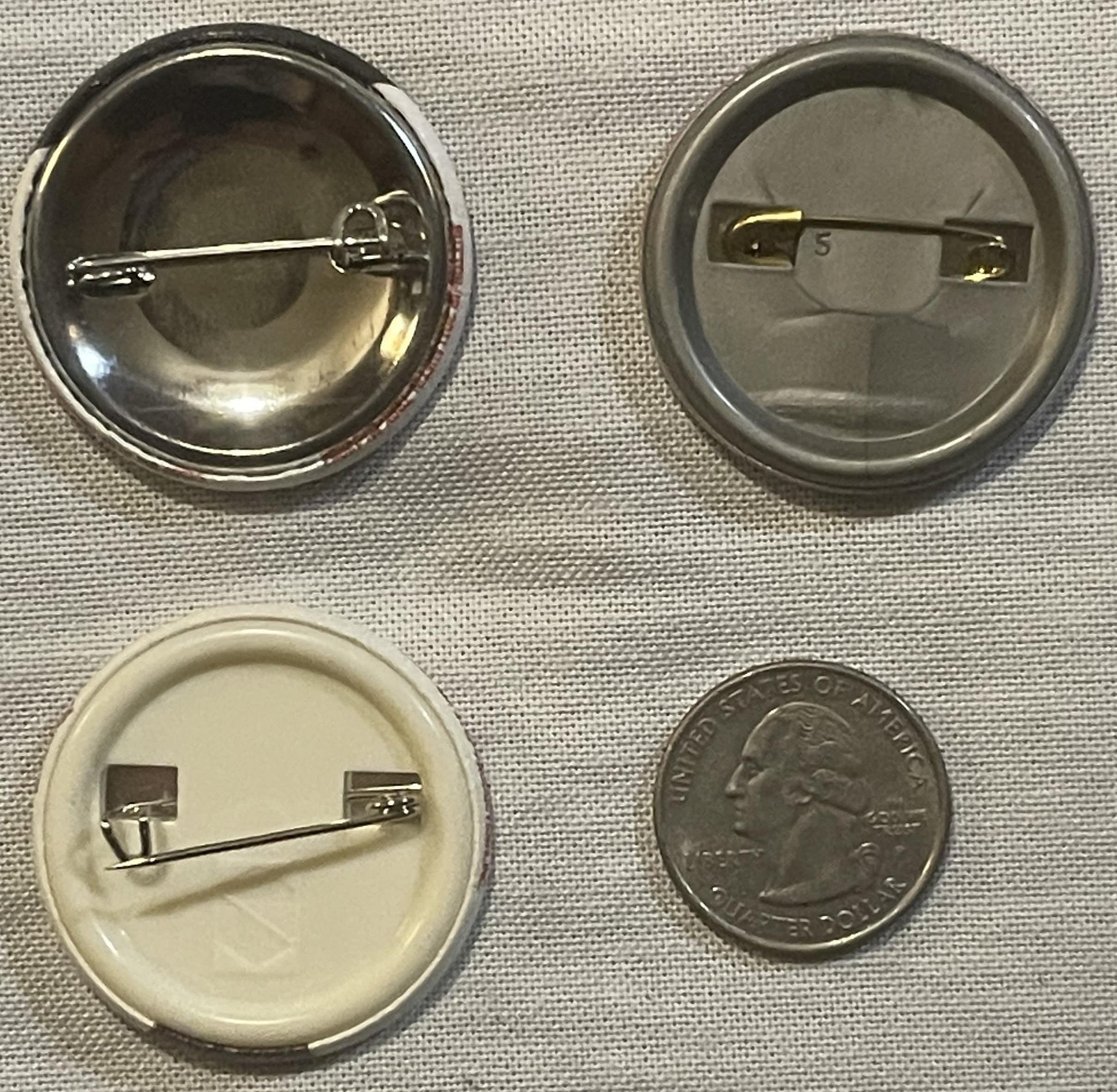 Four pinback buttons shown front and back with a quarter for scale; metal pin backs visible on reverse sides, one button appe