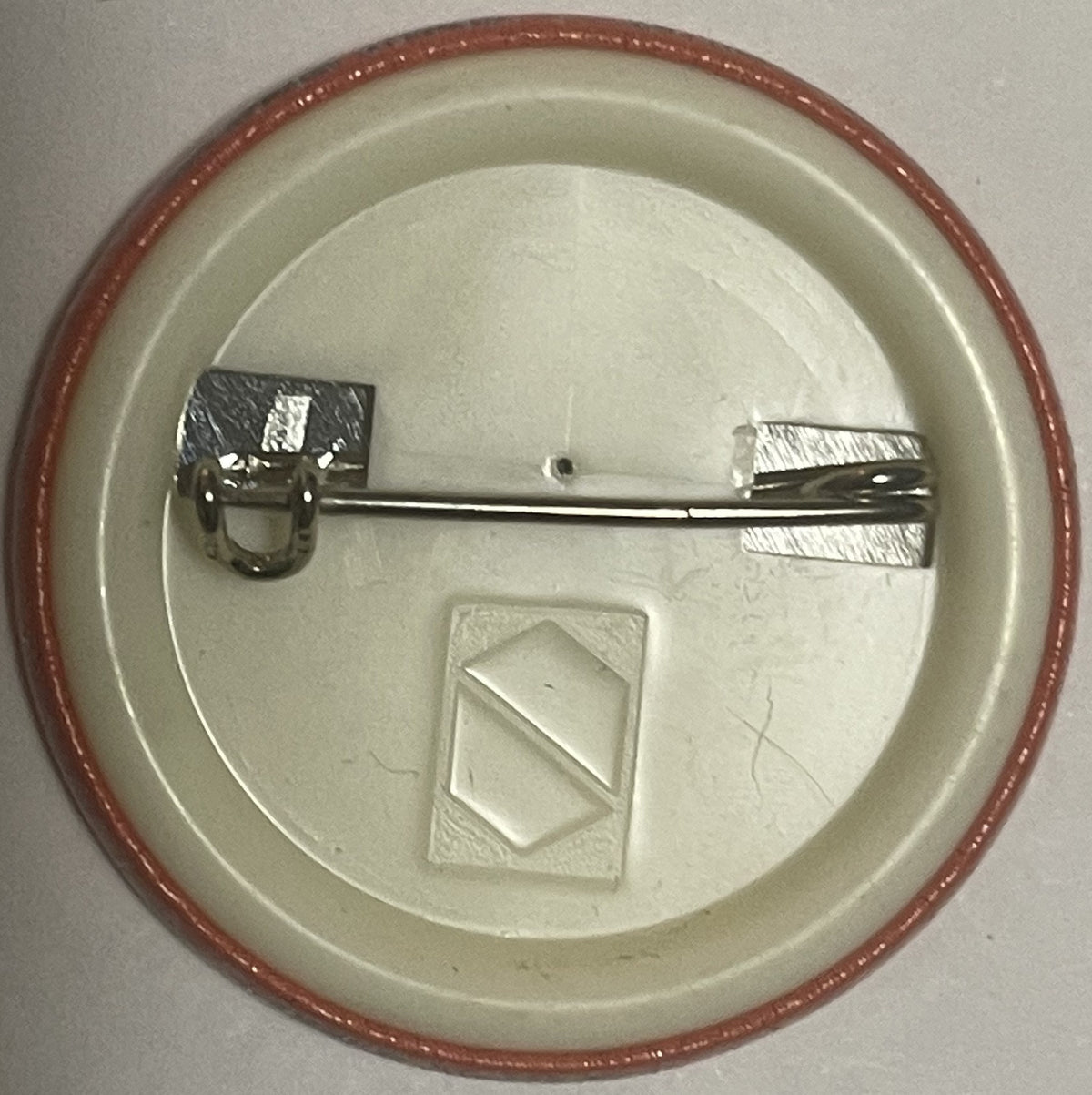 Back of circular pin with reddish rim, cream-colored face, metal clasp mechanism, and embossed diamond shape detail