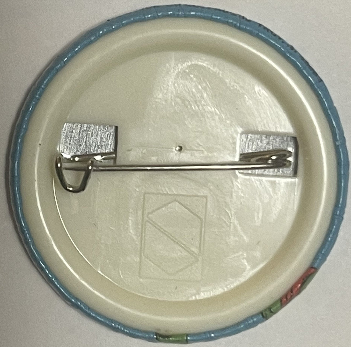 Back of a round pinback button showing metal clasp mechanism and cream-colored plastic backing with embossed shield logo