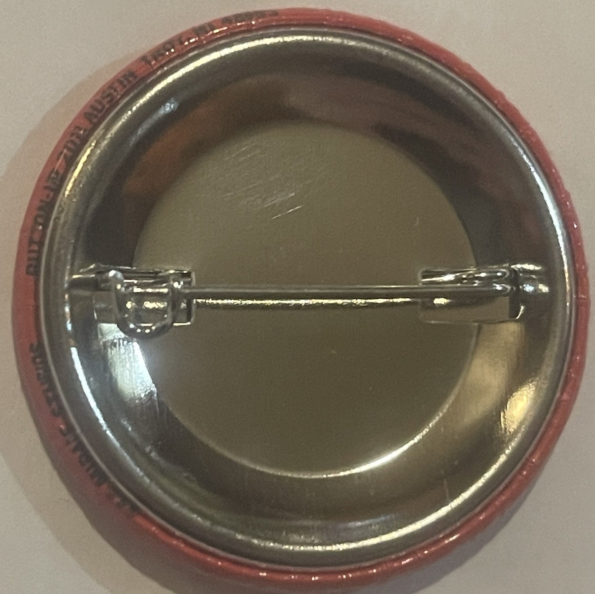 Back of a red and silver oval pinback button with metal pin mechanism visible against beige wall
