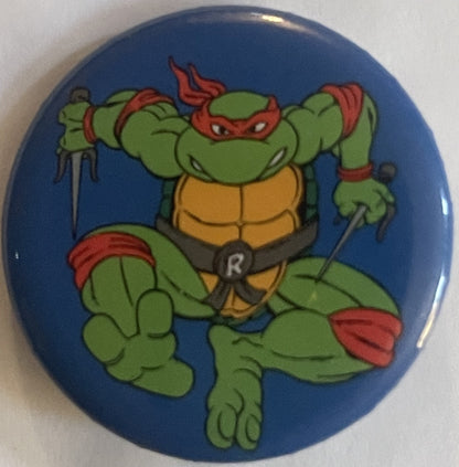Blue circular pinback button featuring a green cartoon turtle with red mask and bandana holding nunchaku, yellow shell, and r