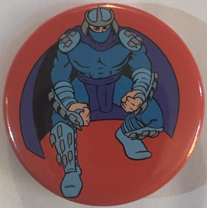 Round red pin badge featuring blue armored villain Shredder in fighting stance with clawed gauntlets and purple cape against 