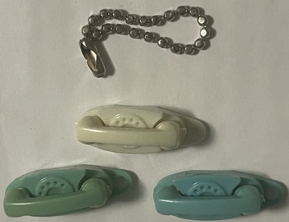 Three miniature rotary telephone keychains in cream, sage green, and teal with metal chain attachment, displayed on white bac