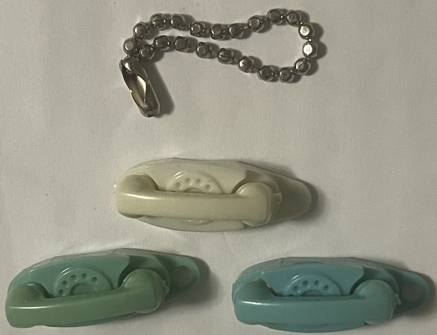 Three miniature rotary telephone keychains in ivory, sage green, and turquoise with metal chain attachment, displayed on whit