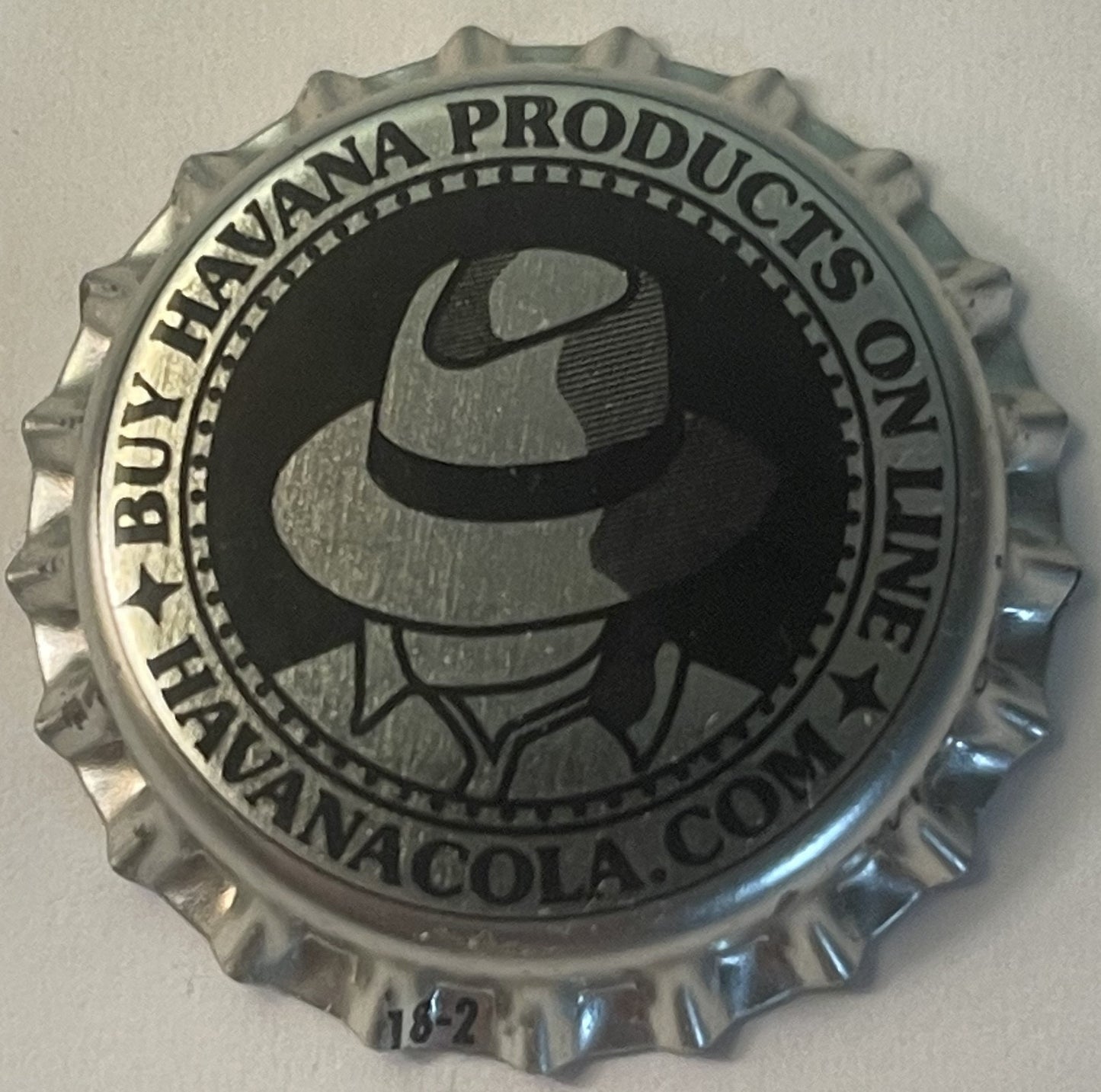 Metallic bottle cap with ridged edges featuring silhouette of man in fedora hat and "Buy Havana Products on Havana Cola, Pens