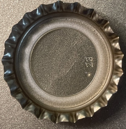 Overhead view of a ridged metal bottle cap with textured center surface and small maker's mark visible, on gray background
