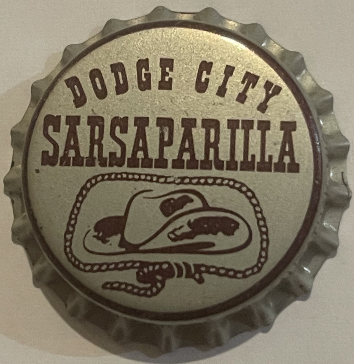 Vintage bottle cap with corrugated metal rim featuring "Dodge City Sarsaparilla" text and illustrated cowboy hat design