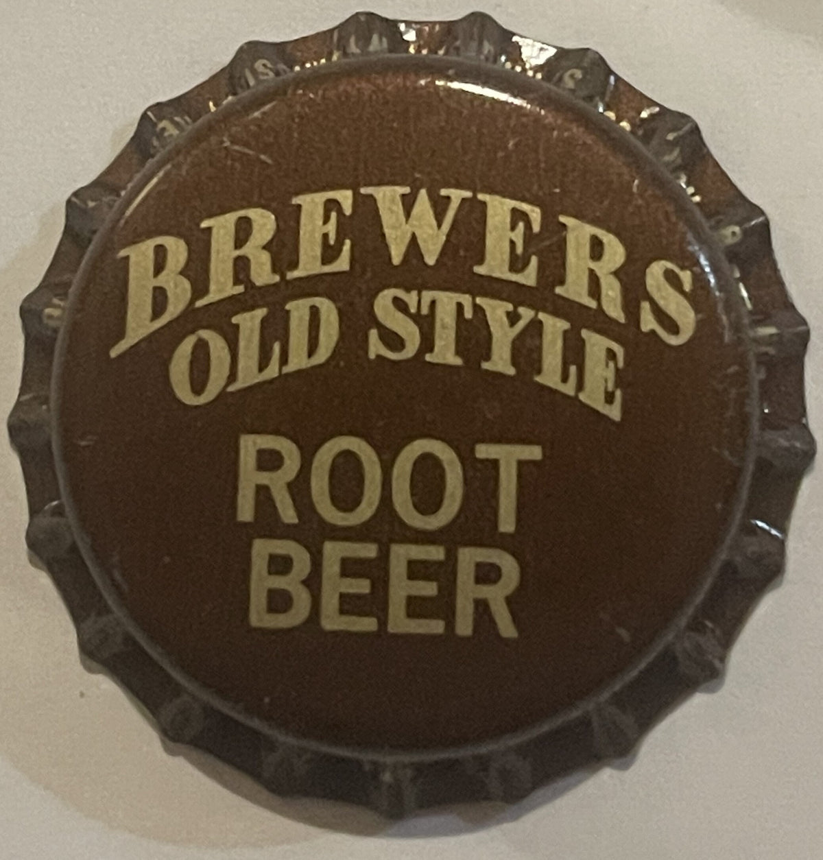 Brown bottle cap with scalloped edges and tan text reading "BREWERS OLD STYLE ROOT BEER," shown flat against a light backgrou