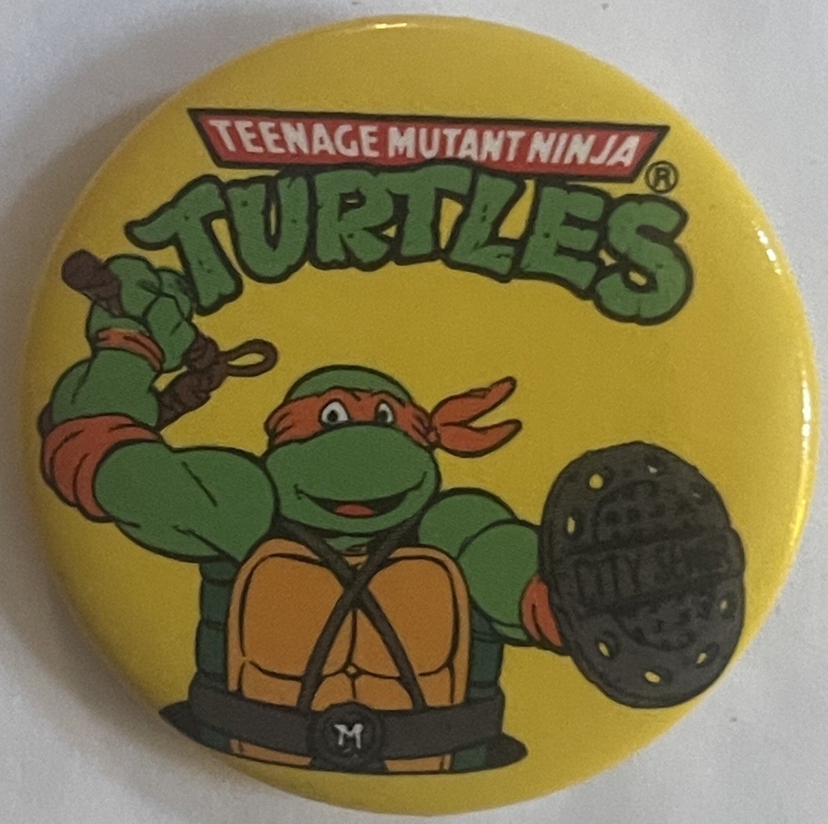 Yellow circular pin featuring Michelangelo from Teenage Mutant Ninja Turtles in action pose with nunchaku, red mask, green