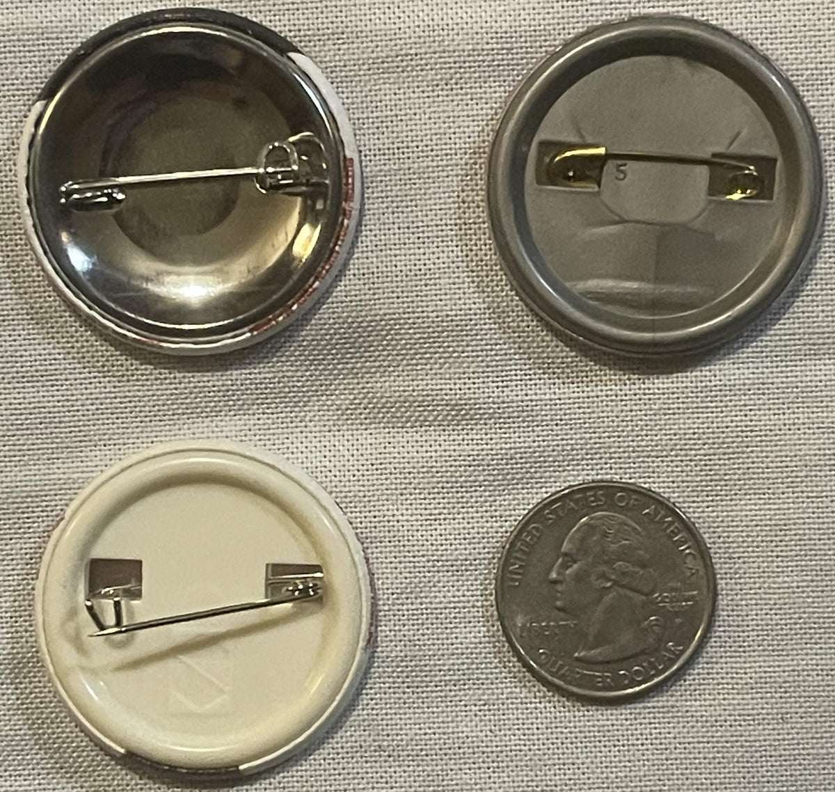 Four button pins shown front and back with a quarter for scale; three metal pins with metallic finishes and one cream-colored