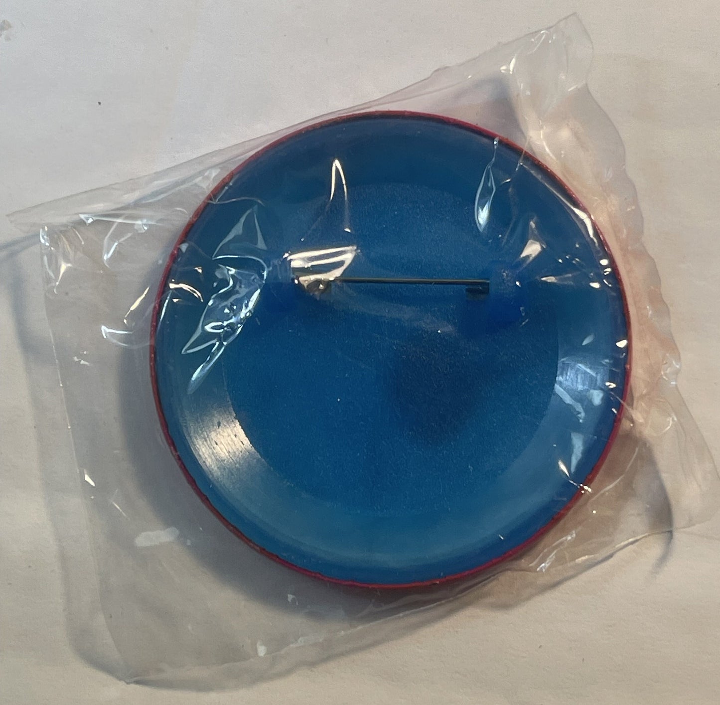 Blue circular enamel pin with red rim in plastic packaging, featuring white sailboat design and black hour hand, displayed on