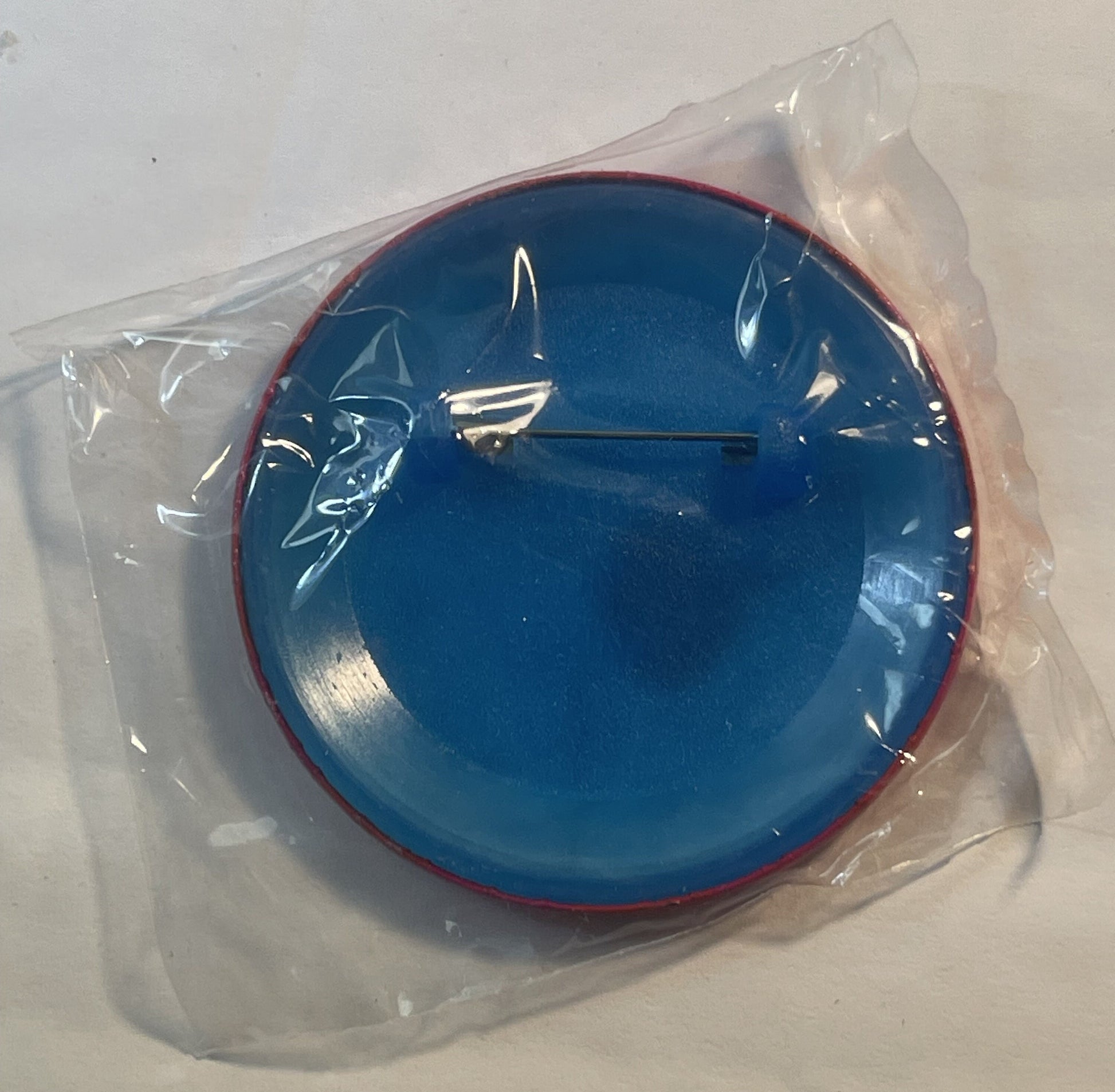 Blue circular enamel pin with red rim in plastic packaging, featuring white sailboat design and black hour hand, displayed on
