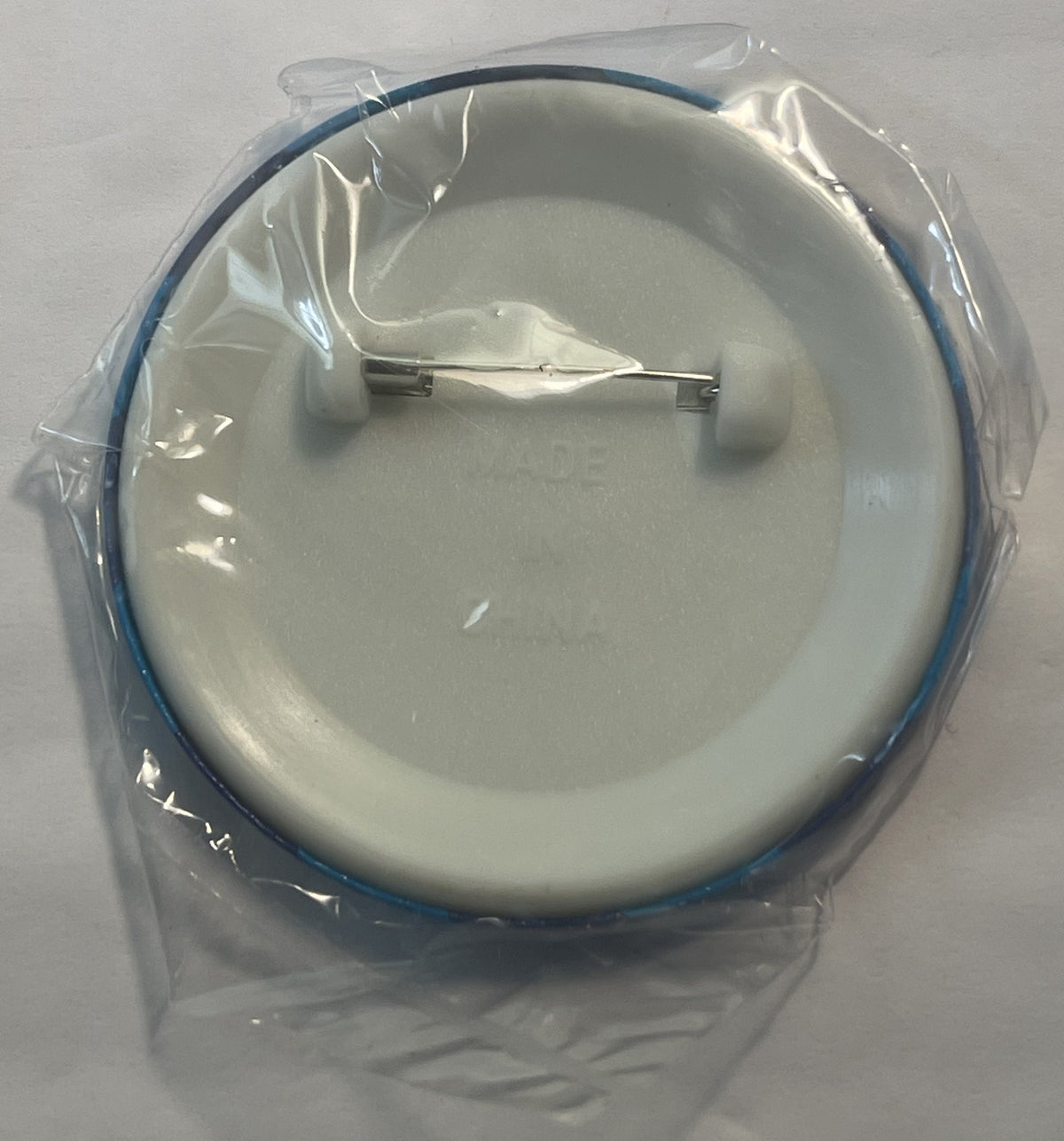 Sealed clear plastic package containing a round white ceramic pin with blue rim and metal clasp, viewed from above