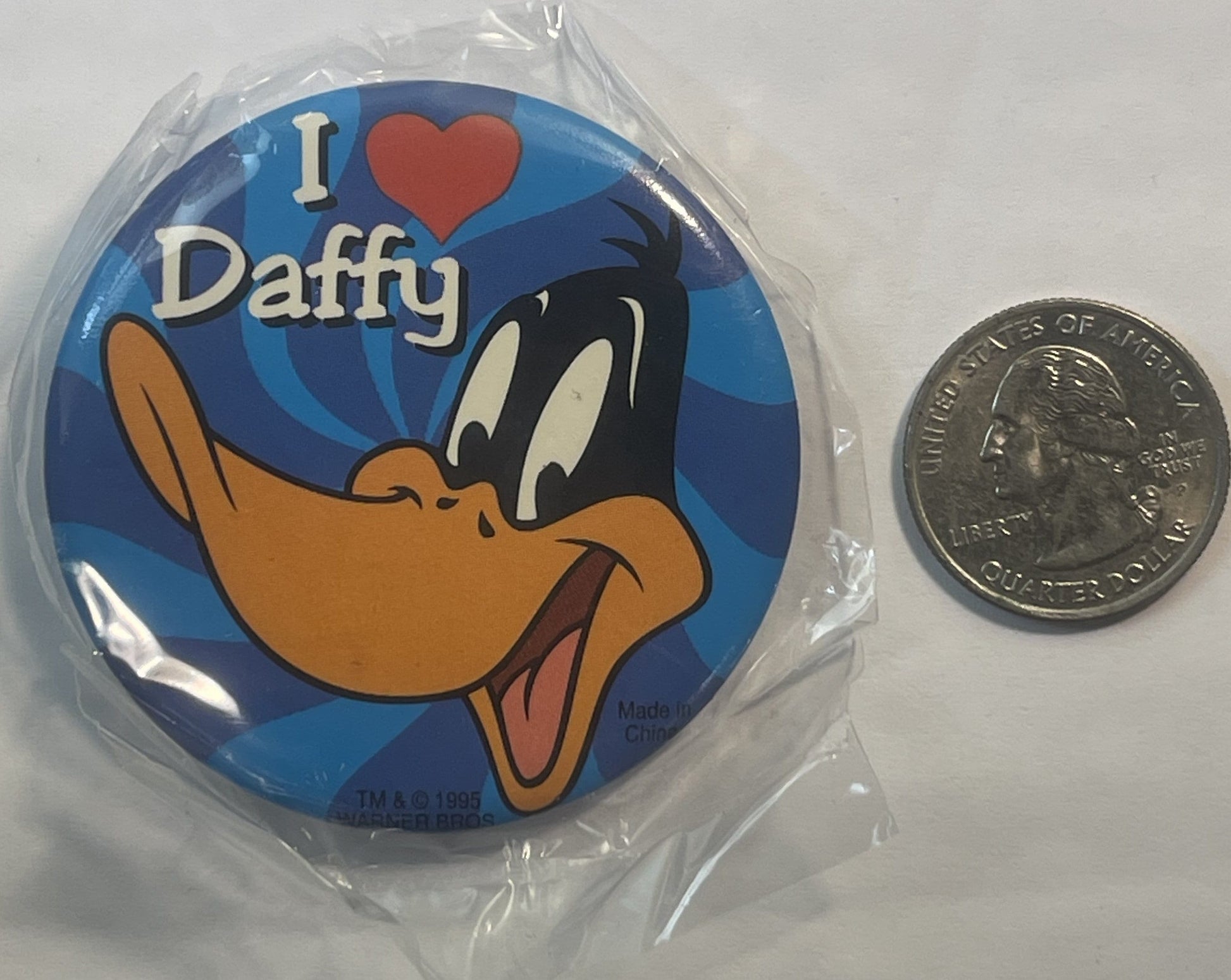 Unopened 1995 Daffy Duck pin in plastic wrapper next to a quarter for scale, showing cartoon duck with "I ♥ Daffy" text on bl