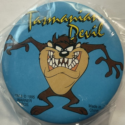 Circular blue pin featuring Tasmanian Devil cartoon character with bared teeth, brown fur, tan face, enclosed in clear plasti