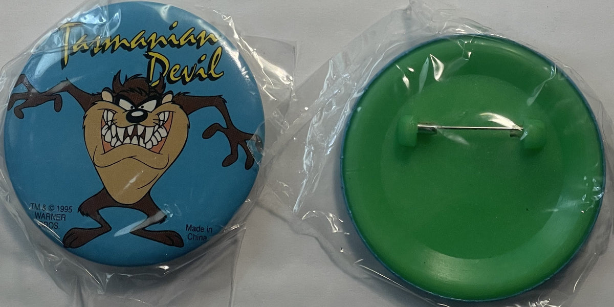 Two circular pins in plastic packaging: blue pin featuring Tasmanian Devil cartoon character with "TM © 1995 Warner" text, an