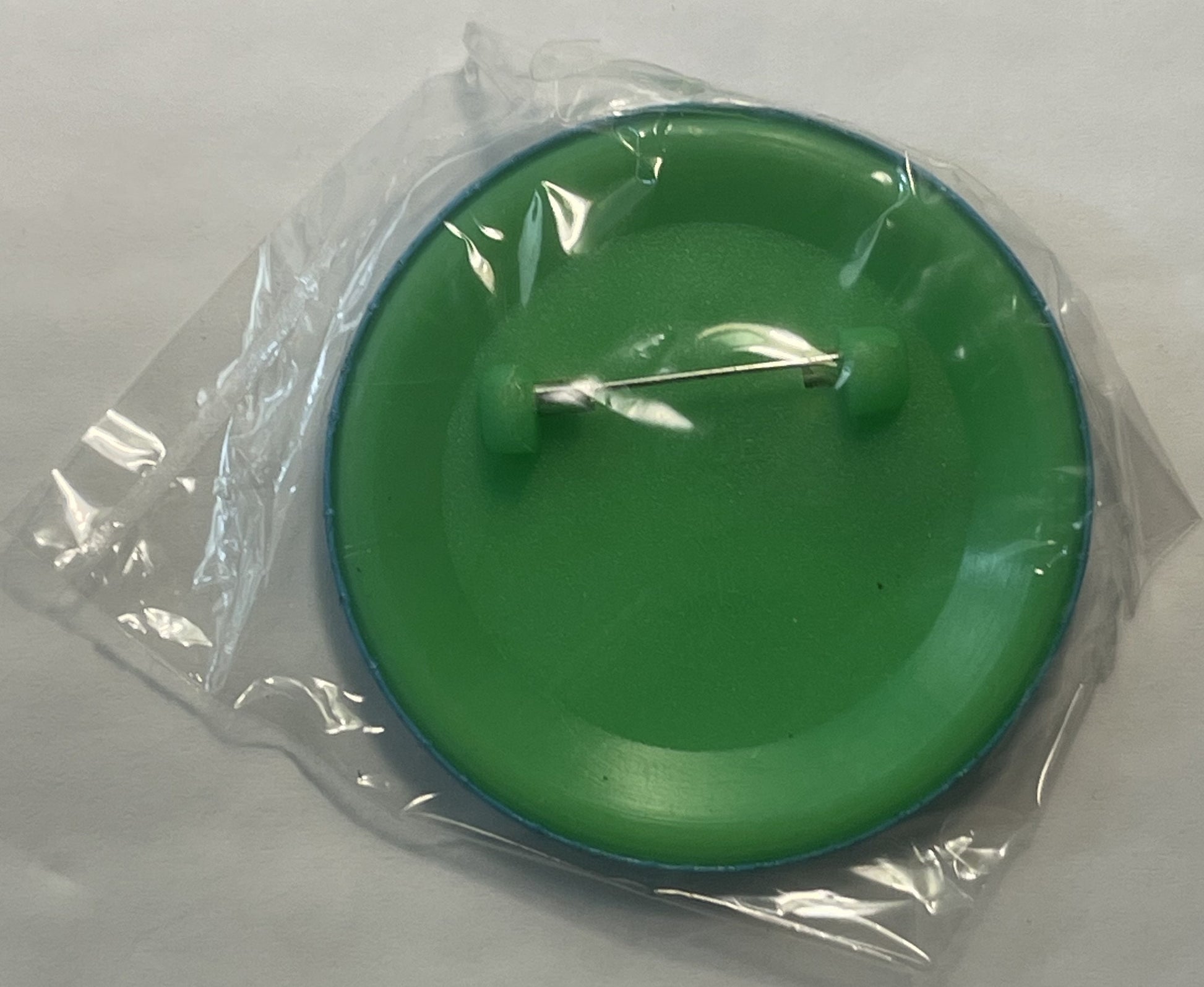 Green circular pin with silver Tasmanian Devil design, still in clear plastic packaging, shown flat against white surface