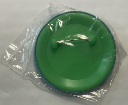 Green circular pin with silver Tasmanian Devil design, still in clear plastic packaging, shown flat against white surface