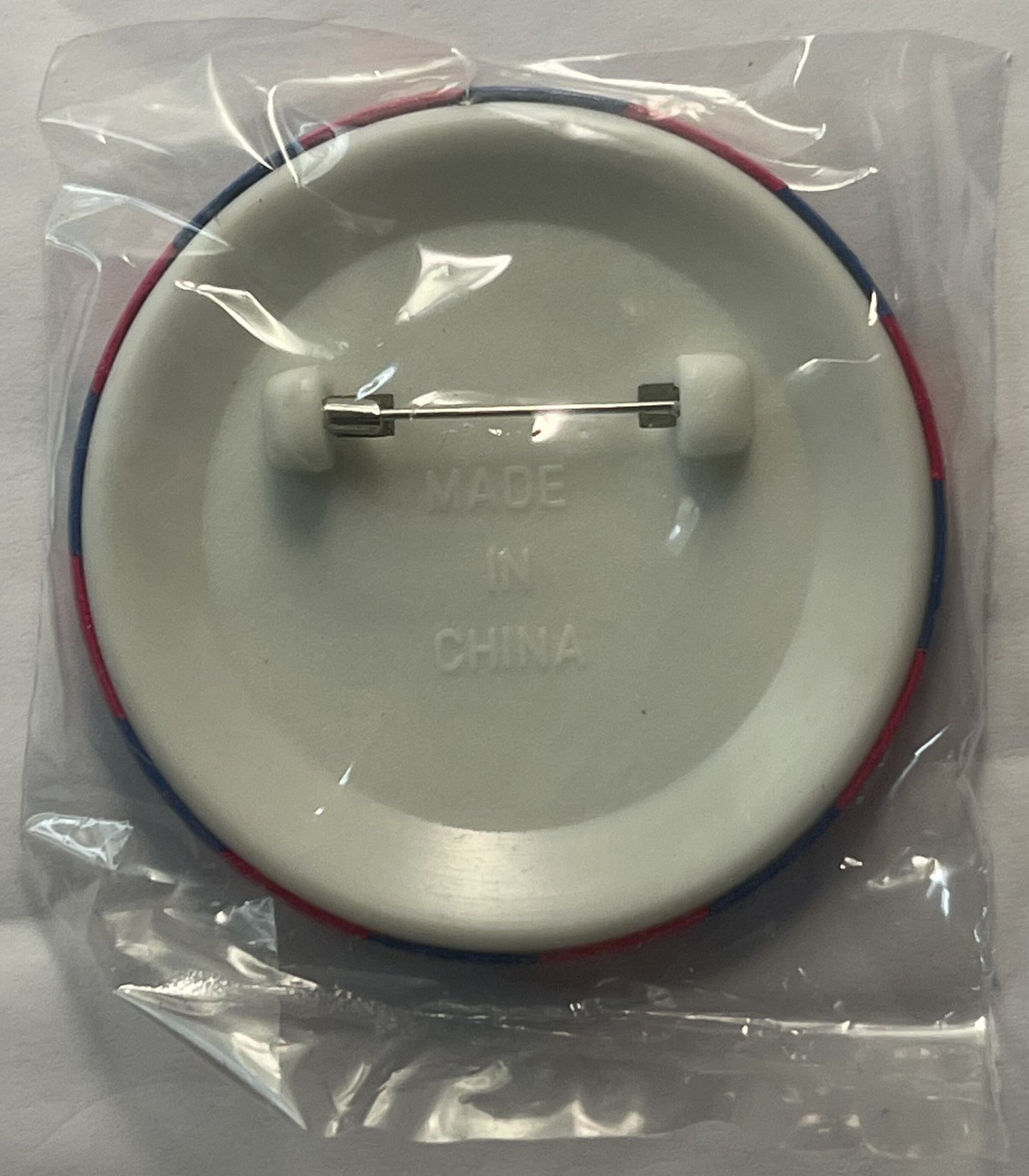 Cream-colored circular pin badge in clear plastic packaging with red and blue fabric straps, metal fastener visible, text emb