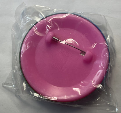 Pink circular pin in clear plastic packaging, viewed from above, with dark rim and metal fastener visible on surface