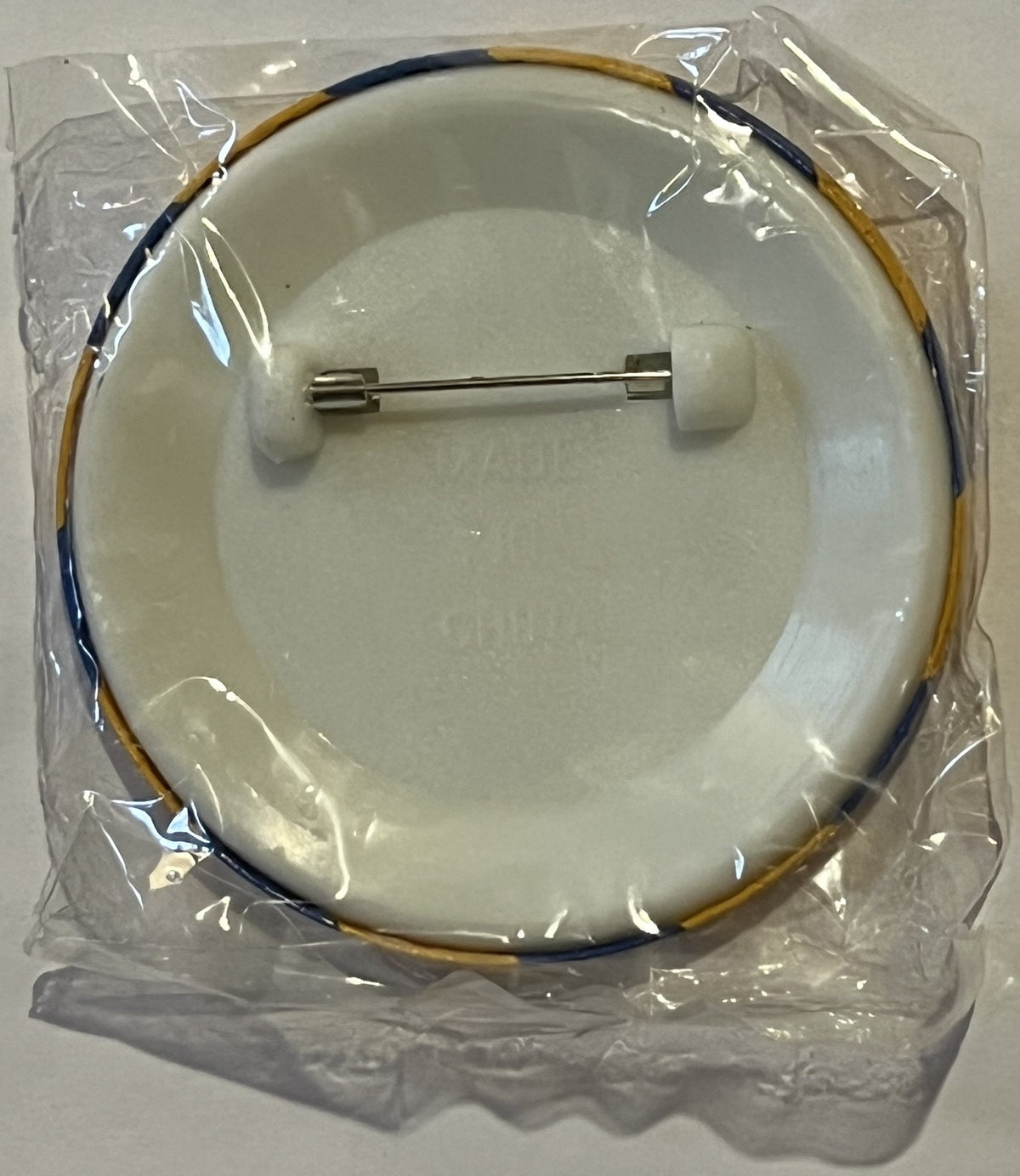 Circular ceramic pin with dark blue and tan cord loop, metal backing pin, sealed in clear plastic packaging