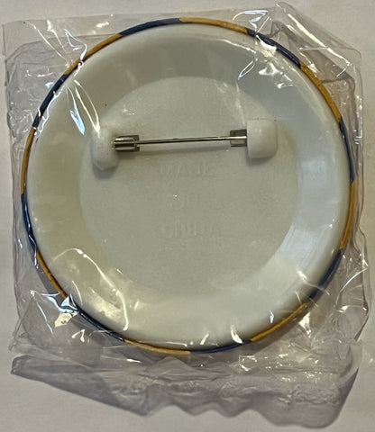 Circular ceramic pin with dark blue and tan cord loop, metal backing pin, sealed in clear plastic packaging