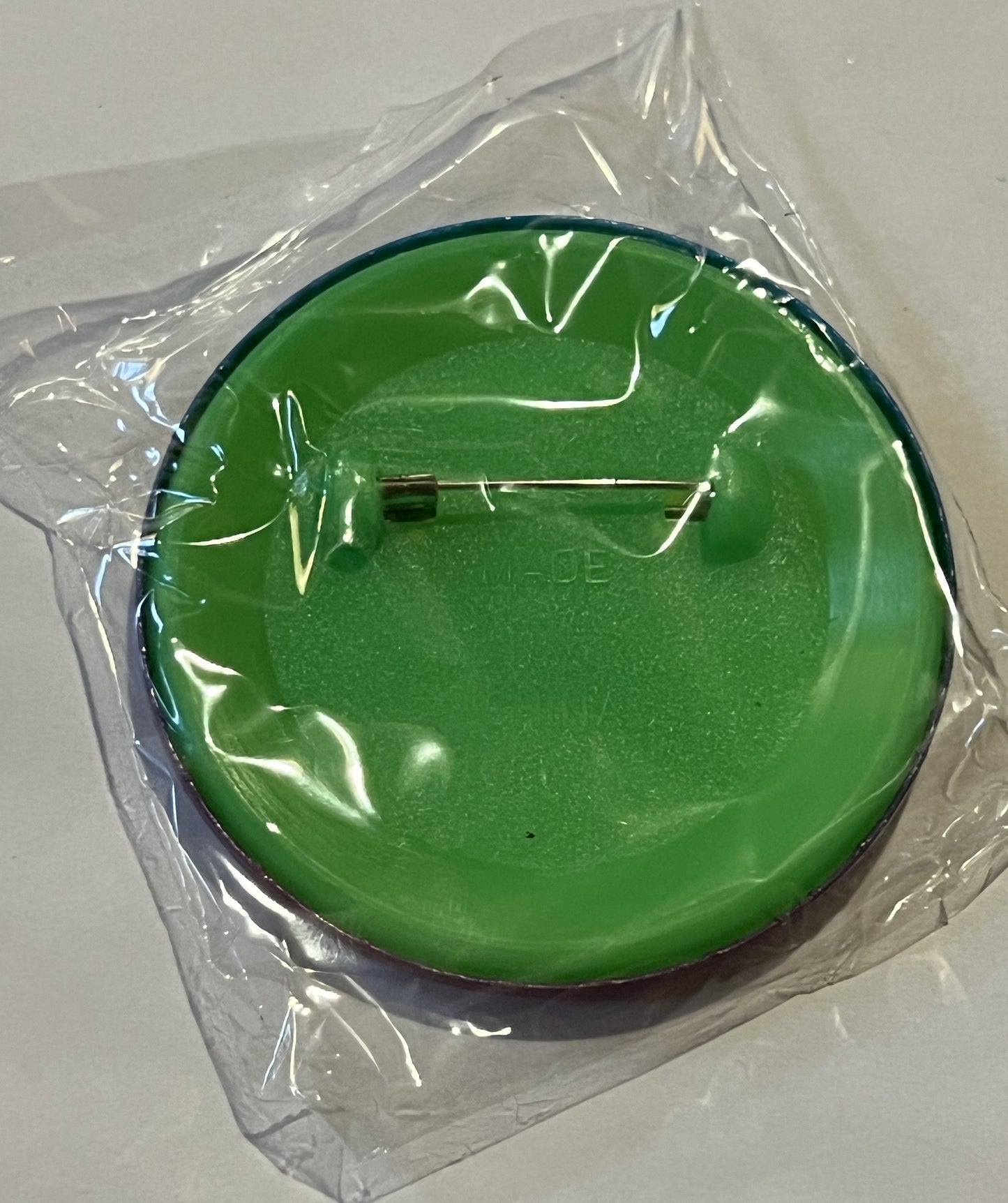 Sealed clear plastic package containing a bright green circular pin with metal clasp, viewed from above against white surface