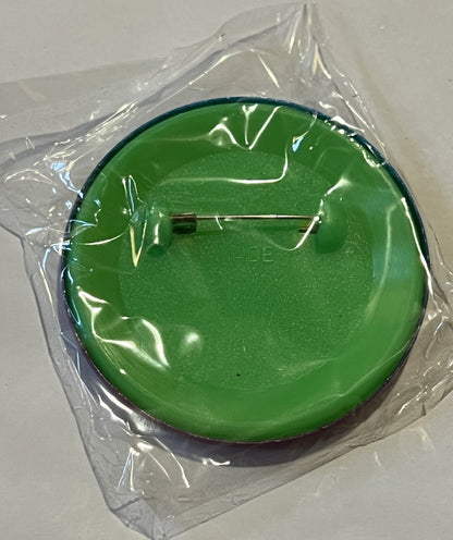 Sealed clear plastic package containing a bright green circular pin with metal clasp, viewed from above against white surface
