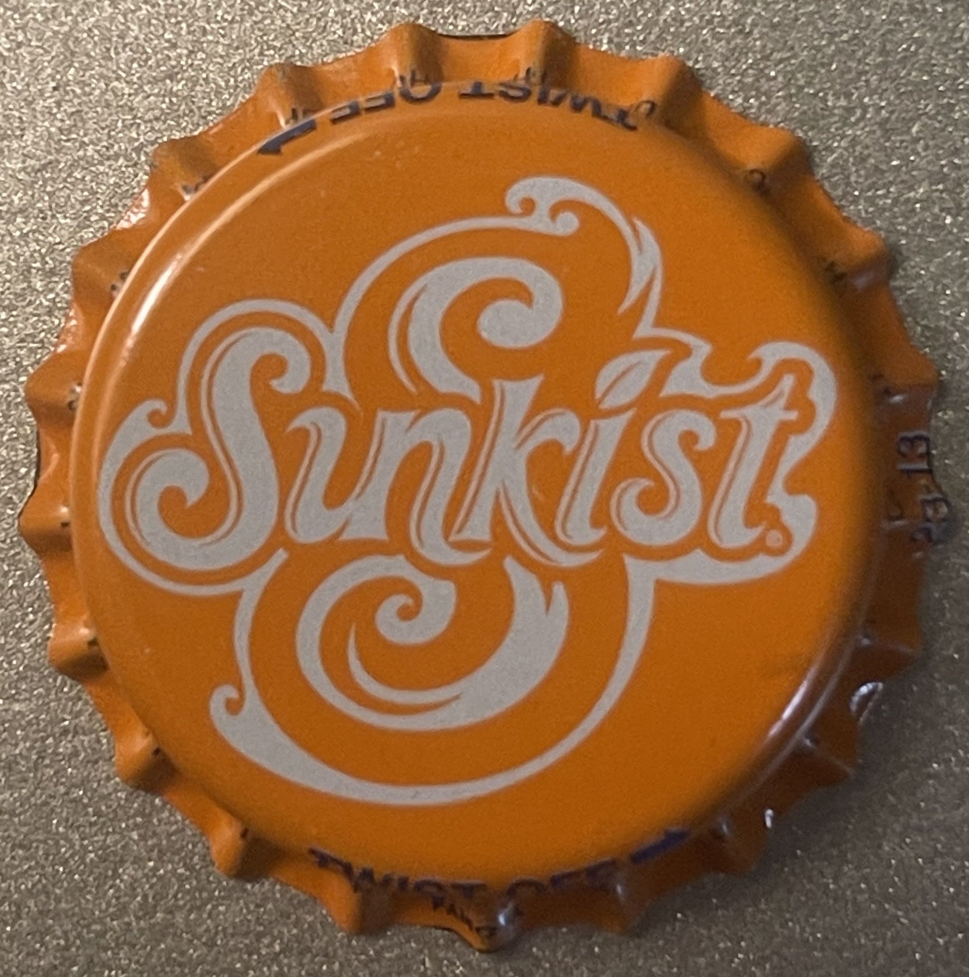 Orange Sunkist bottle cap with white decorative scrollwork logo, shown flat against textured gray surface