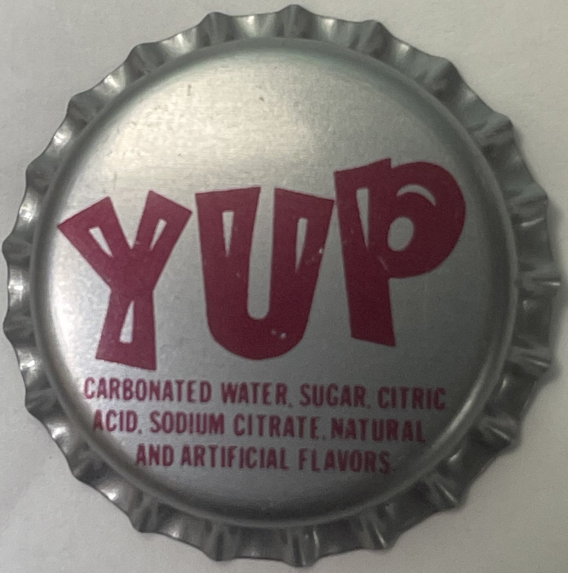 Vintage metal bottle cap with crimped edges, white center and red "YUP" lettering with ingredients listed below, shown flat a