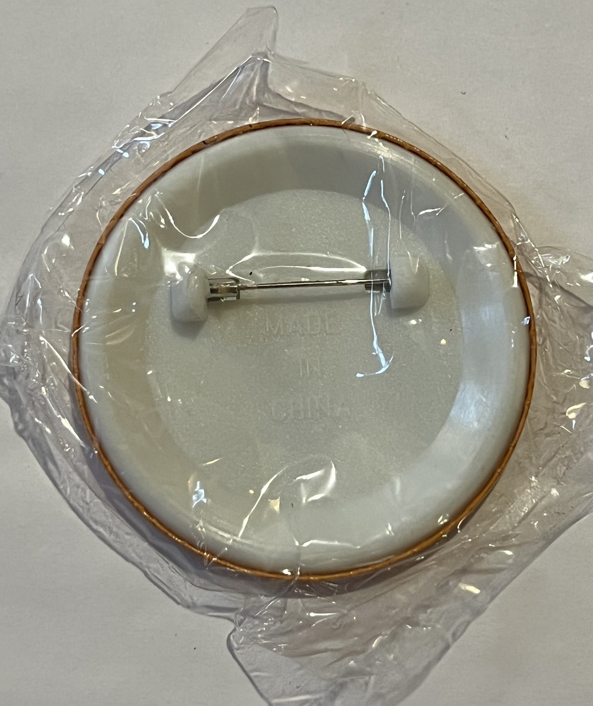 Cream-colored ceramic plate with brown rim in clear plastic packaging, showing metal pin backing, viewed from above