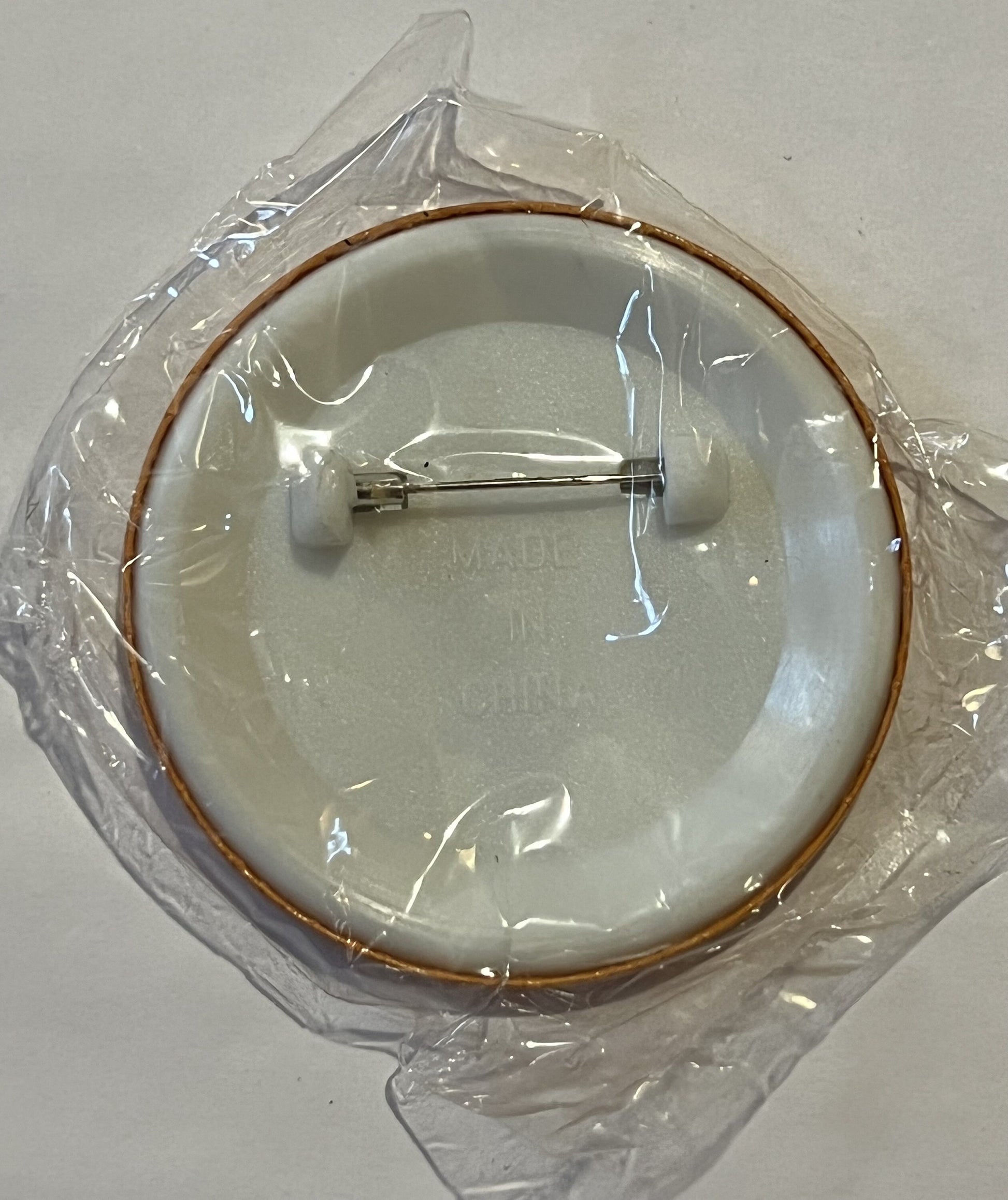 Cream-colored ceramic plate with brown rim in clear plastic packaging, showing metal pin backing, viewed from above