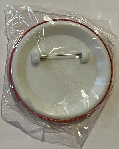 Unopened circular pin badge in clear plastic packaging with red trim, featuring white button back and metal pin, viewed from 