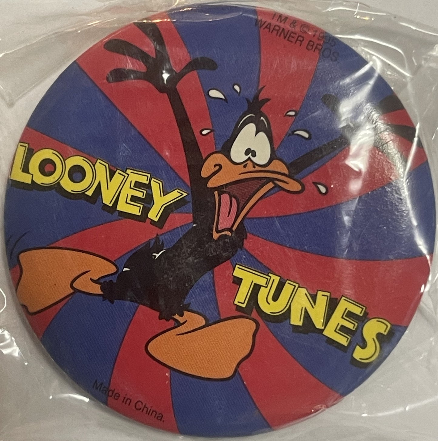 Circular pin featuring cartoon Daffy Duck with yellow "LOONEY TUNES" text on red and blue radial background, sealed in clear 