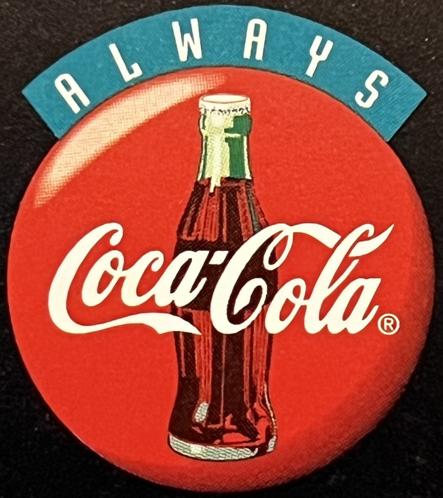 Circular red Coca-Cola door hanger with bottle image, "ALWAYS" text on teal banner at top, displayed against dark textile bac