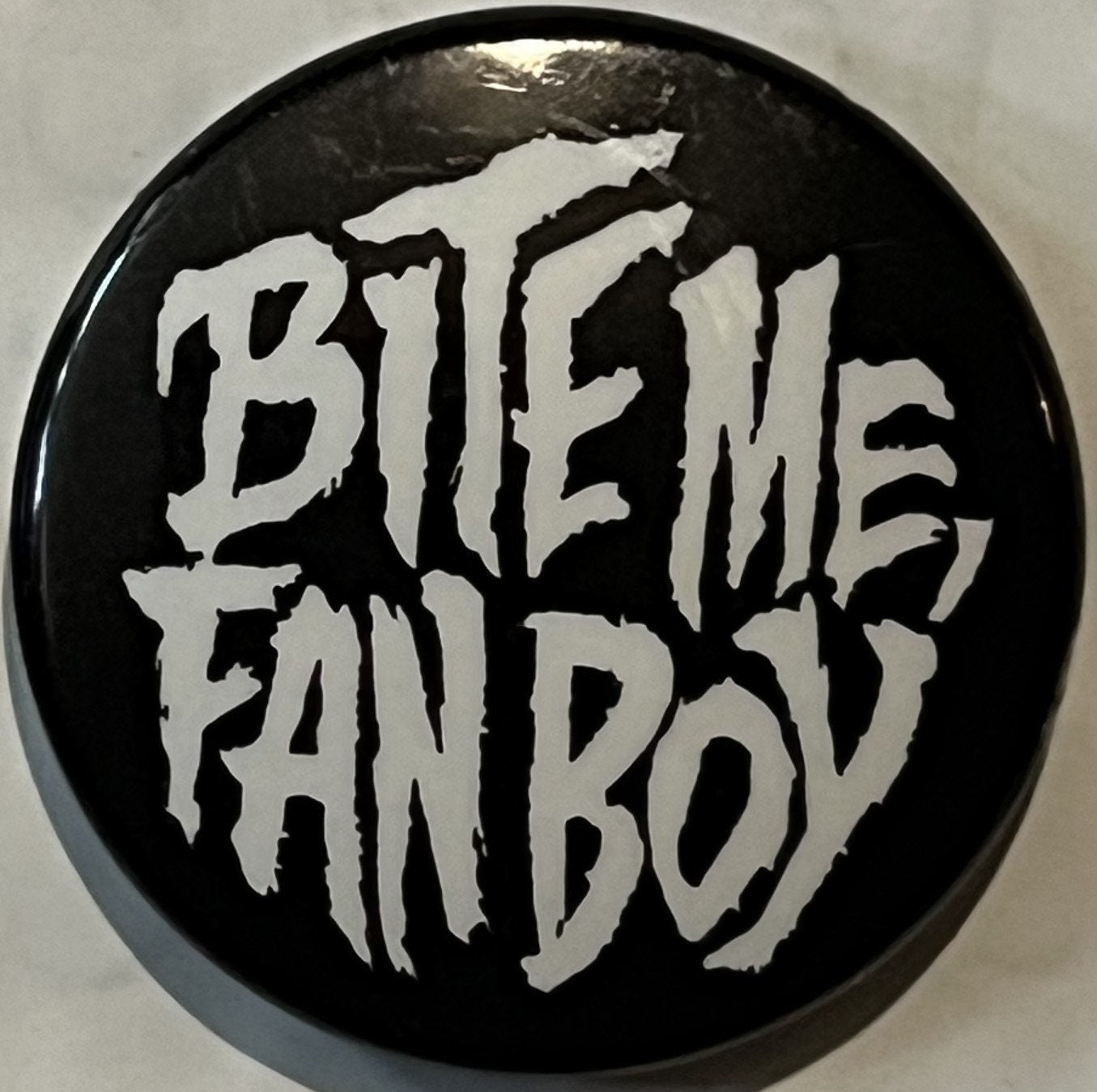 Black circular pinback button with white distressed lettering reading "BITE ME FAN BOY" against light background