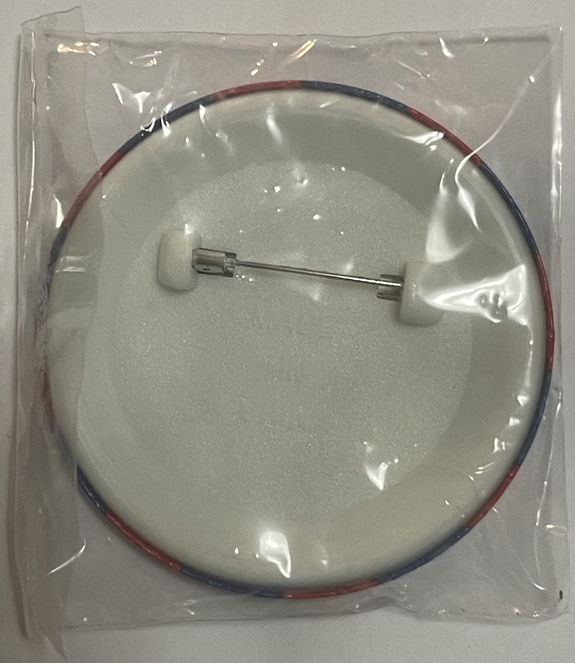 Sealed plastic package containing a circular pin with red and blue wire attachment and metal clasp, mounted on white foam bac