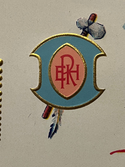 Gold-bordered oval emblem with turquoise background and coral center containing red initials, flanked by decorative feathers,