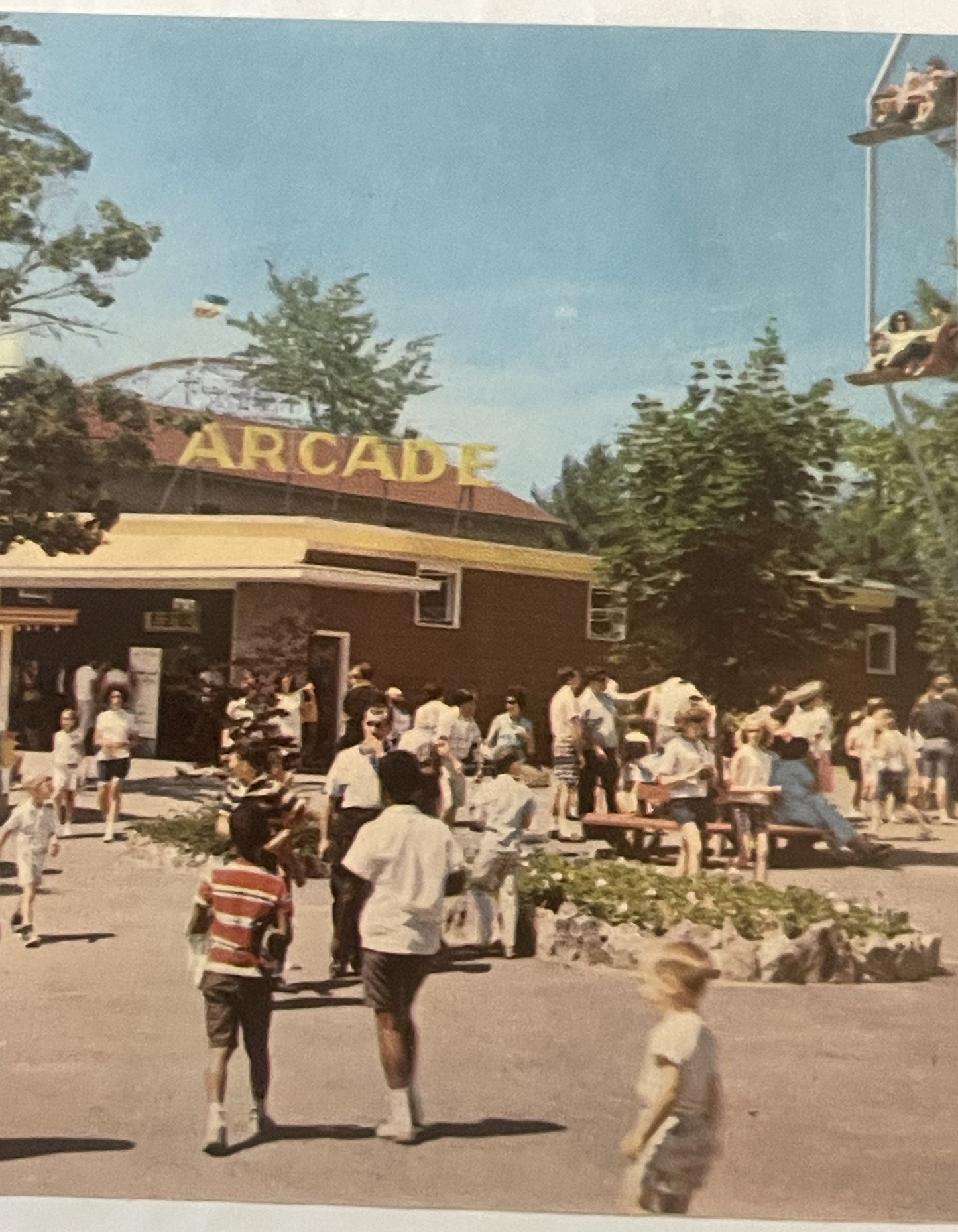 Vintage arcade building with yellow lettering surrounded by visitors in 1960s summer clothing, with evergreen trees and amuse