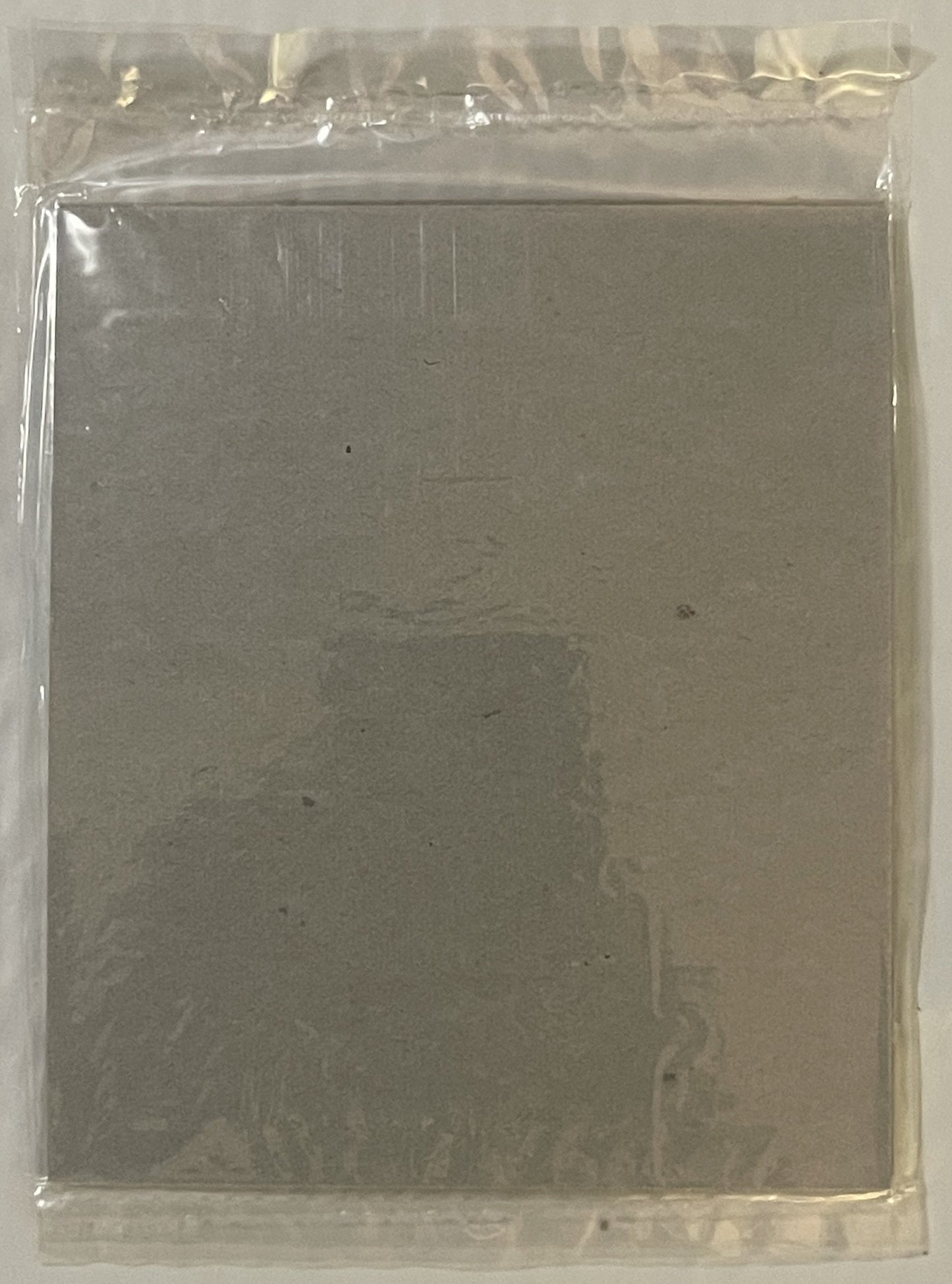 Sealed plastic package containing a grey cardboard puzzle box with faint printed text visible through the packaging