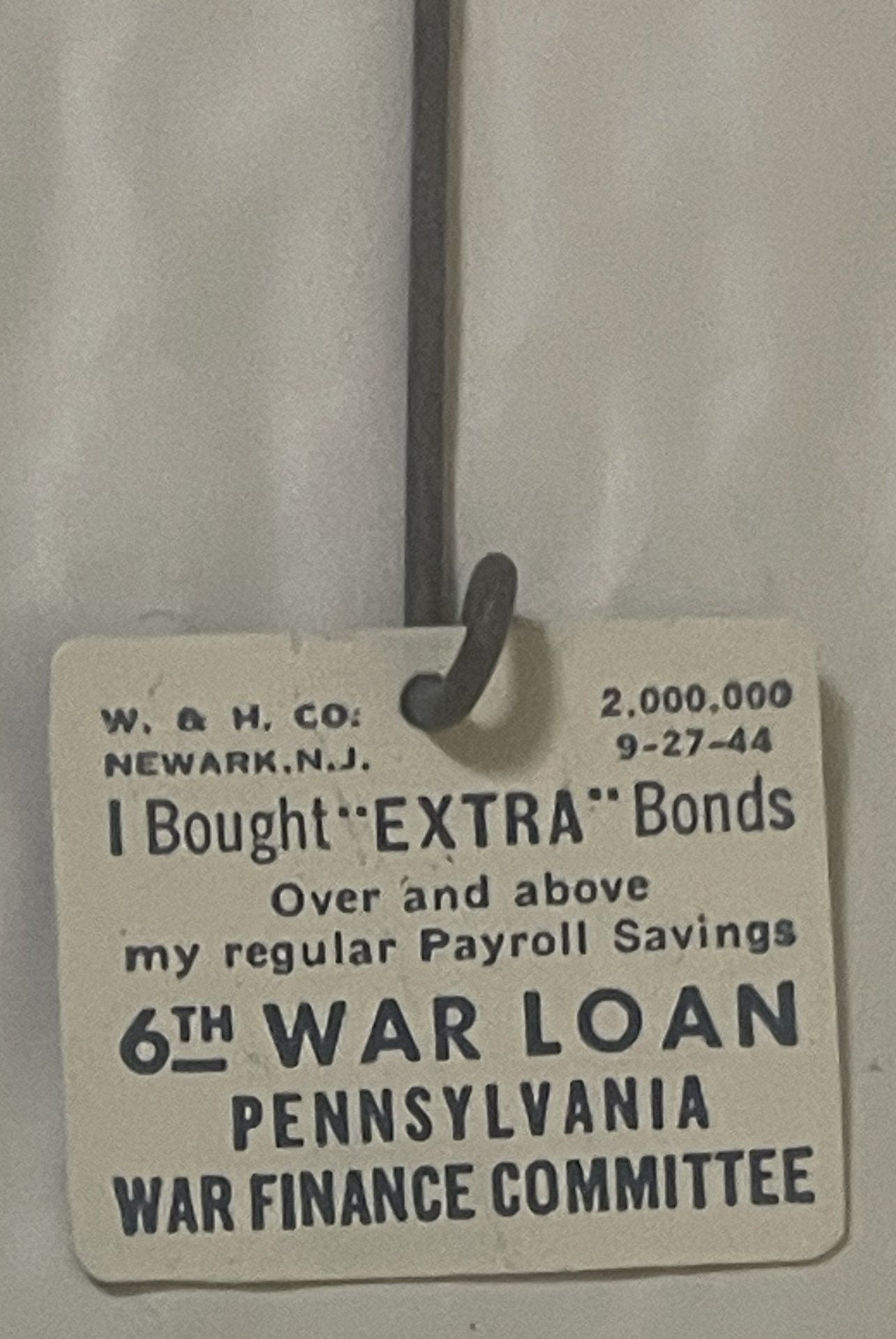Cream-colored WWII war bond badge from Newark, N.J., dated 9-27-44, hanging from black metal clasp against gray curtain backd