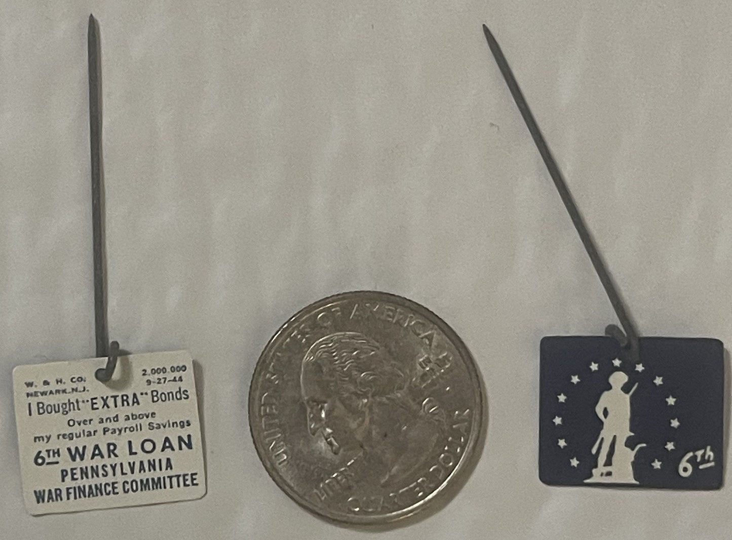 Three WWII-era badges displayed with a quarter for scale: cream tag reading "6TH WAR LOAN PENNSYLVANIA" on left, silver coin 