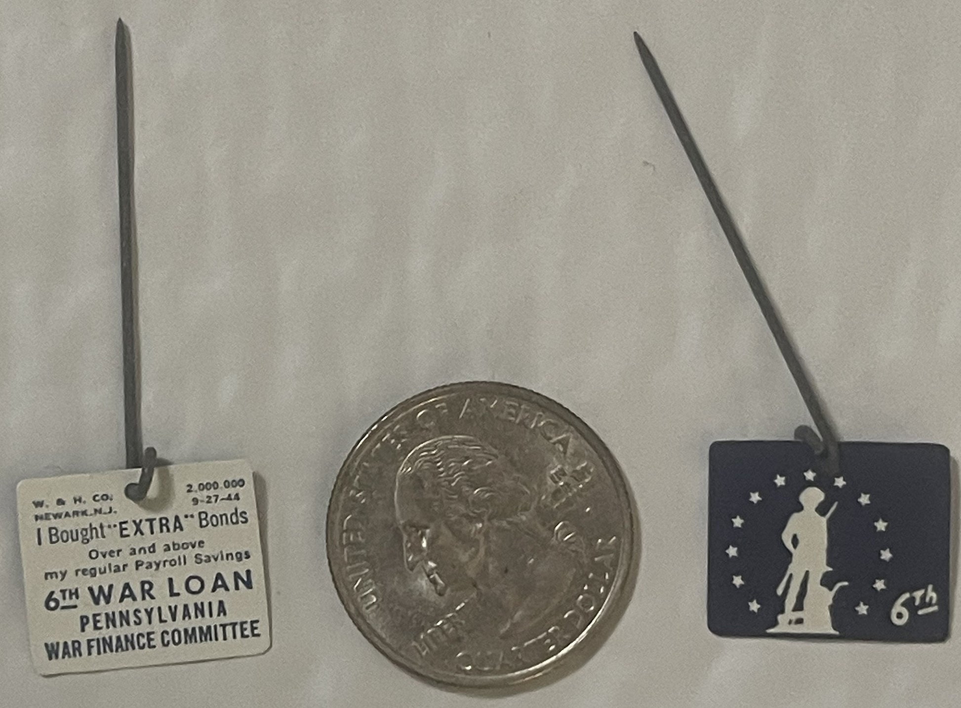 Three WWII-era badges displayed with a quarter for scale: cream tag reading "6TH WAR LOAN PENNSYLVANIA" on left, silver coin 