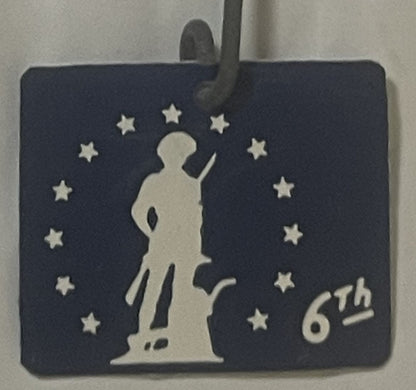 Dark blue badge with cream minuteman silhouette surrounded by stars and "6th" text, displayed on holder with thumb visible at