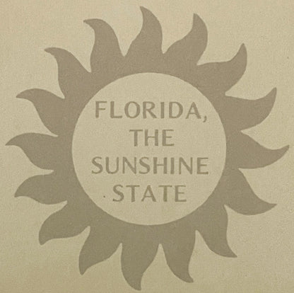 Tan vintage postcard with embossed sunflower design surrounding circular center text reading "FLORIDA, THE SUNSHINE STATE