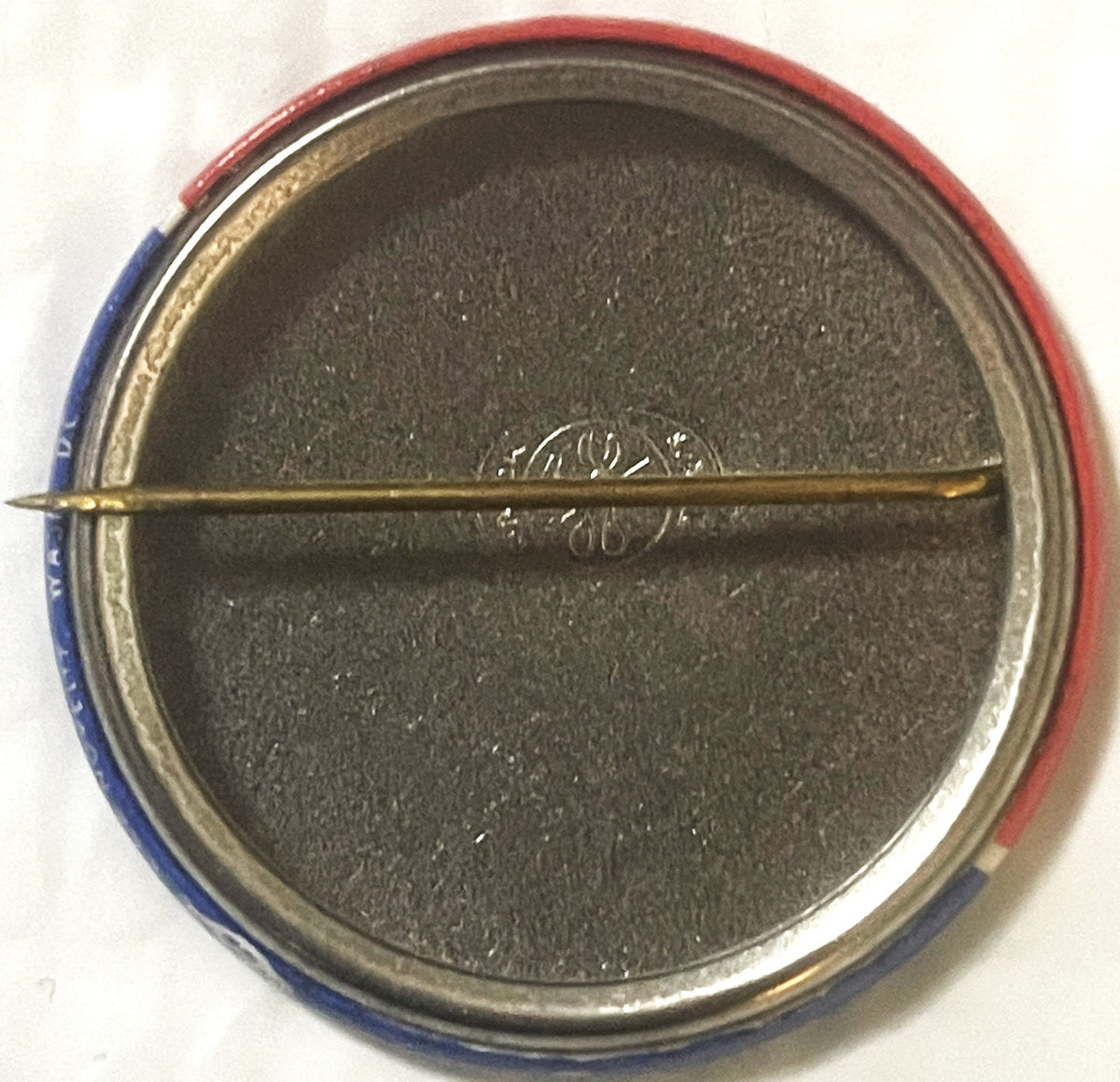 The reverse of the button: a plain metal back with the rolled red and blue edge of the front just visible, and a brass safety pin across the middle.