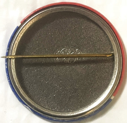 The reverse of the button: a plain metal back with the rolled red and blue edge of the front just visible, and a brass safety pin across the middle.