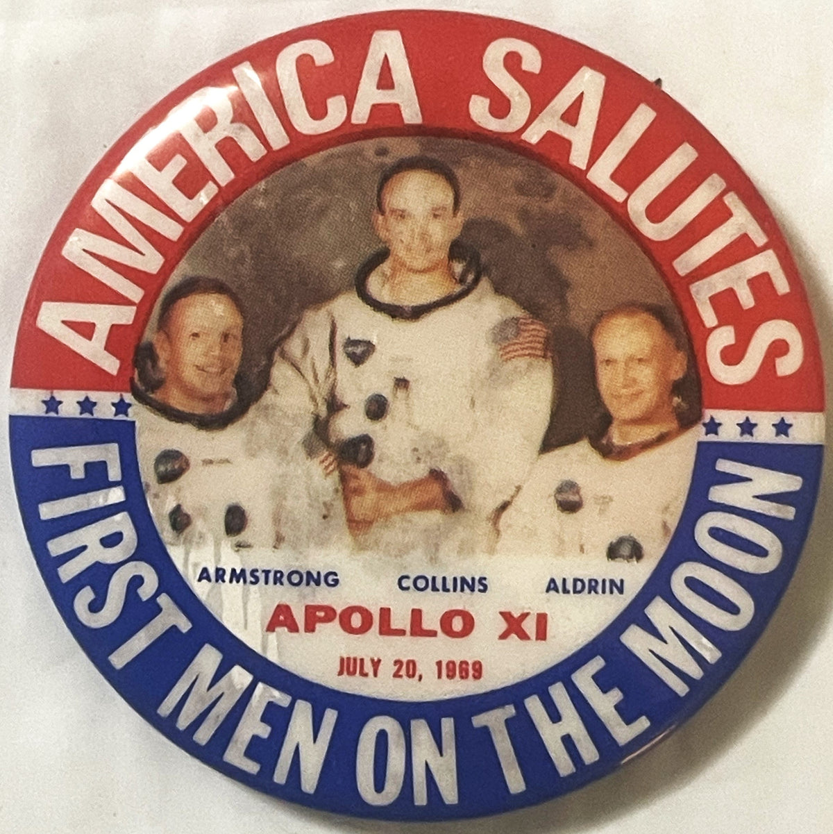 Front of the Apollo 11 pinback button: the NASA crew portrait of Armstrong, Collins and Aldrin in white suits, ringed by AMERICA SALUTES above and FIRST MEN ON THE MOON below in red and blue, with APOLLO XI and JULY 20, 1969 beneath the photograph.