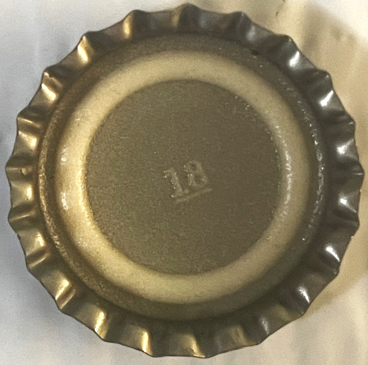 Vintage metal bottle cap with corrugated ridged edges, viewed from above on light background, showing embossed text in center